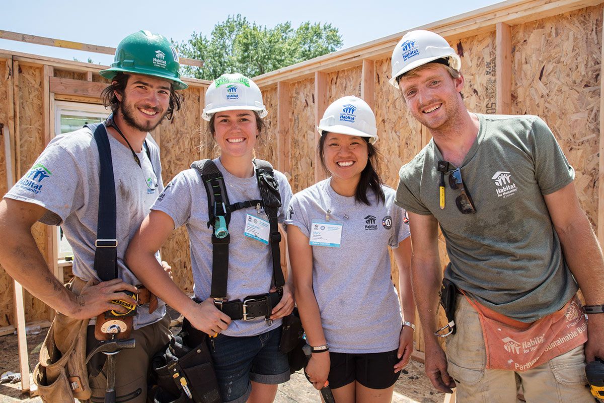 Four young people in hardhats and work clothes at a home build site standing shoulder-to-shoulder and smiling