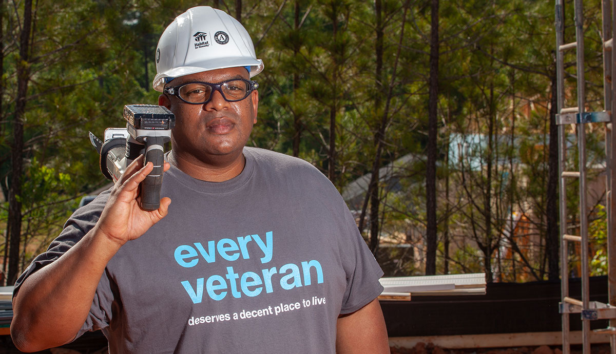 Man in hardhat and t-shirt carrying an electric saw over his shoulder.