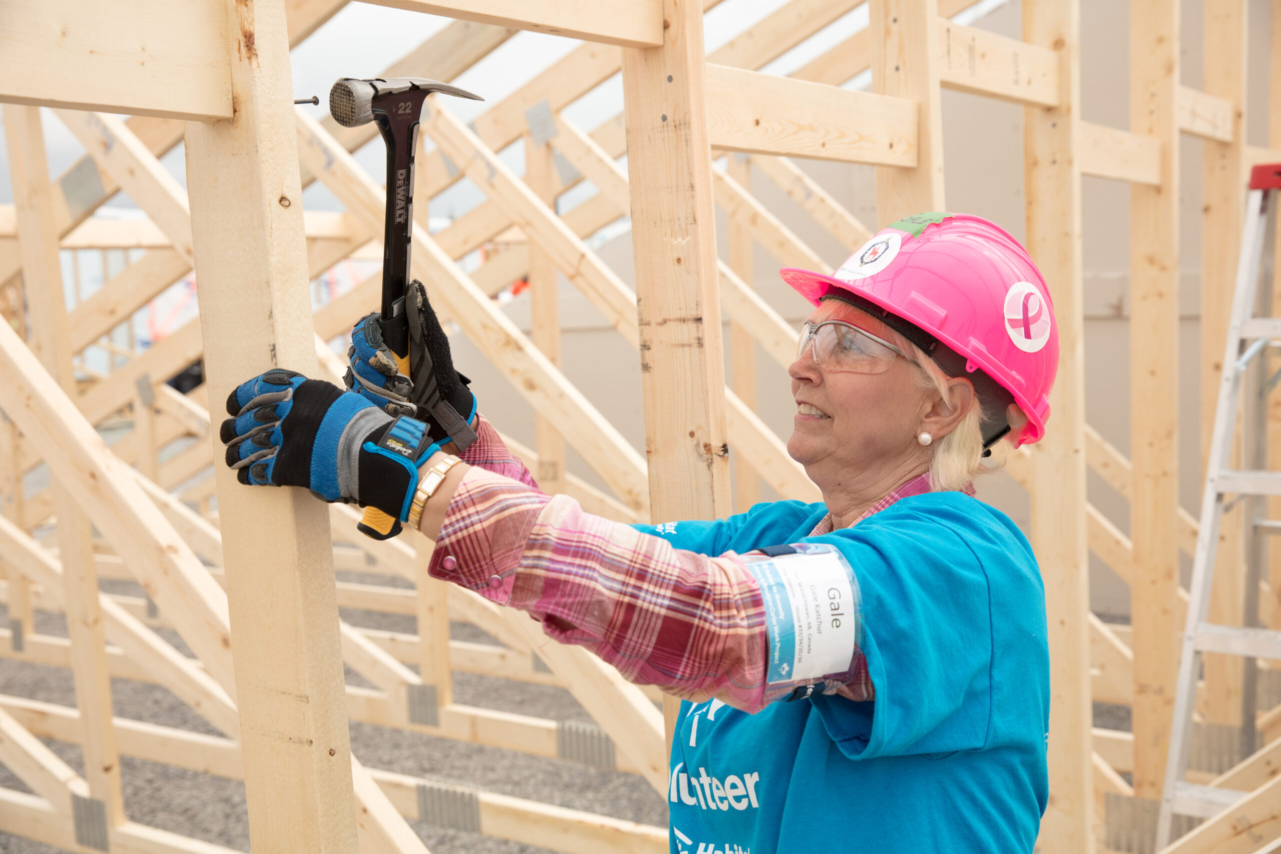 Woman wearing gloves, hardhat, and safety glasses nails wall studs for a home under construction