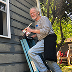A man with gray hair and a short white beard paints the side of a house