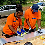 Two homeowner volunteers in orange shirts cut a piece of flashing on a plywood table on a home build site