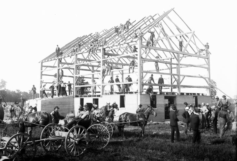 Almost completed barn frame with workers sitting and standing on the frameworks, surrounded by people in wagons, buggies, and standing on the grounds