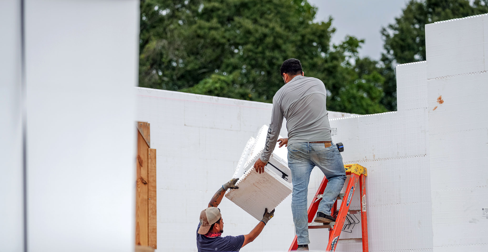 A worker hands an insulating concrete form to another worker who is standing on a ladder by an incomplete wall